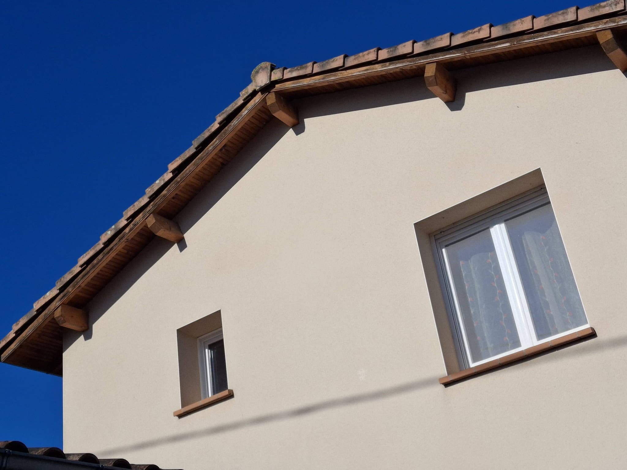 Façade beige avec toit en tuiles, avant-toit en bois et deux fenêtres rectangulaires sous un ciel bleu.