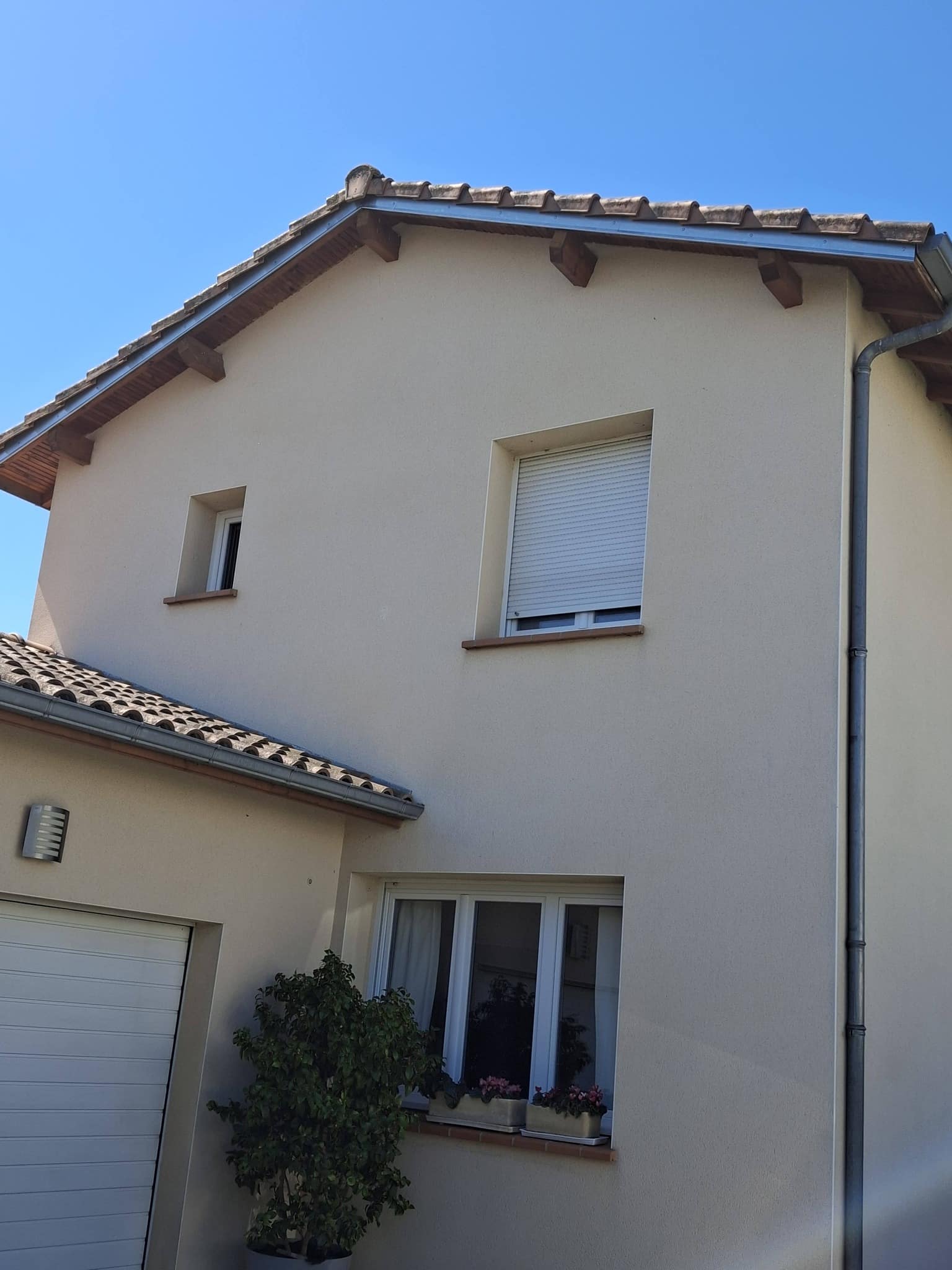 Façade de maison moderne beige avec toit en tuiles, volet roulant et fenêtres blanches sous un ciel bleu ensoleillé.