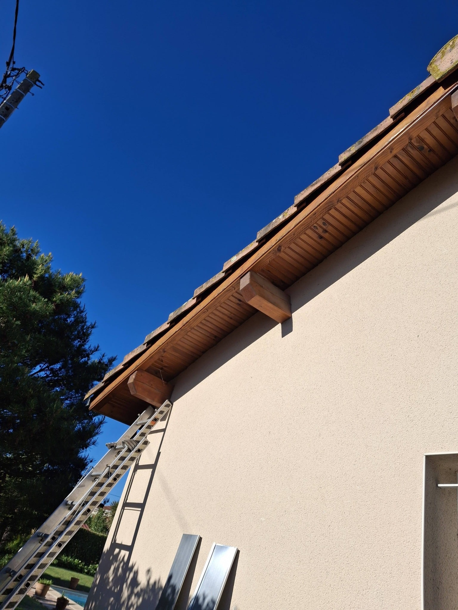 Échelle contre une façade beige sous un débord de toit en bois massif lors de travaux de rénovation.