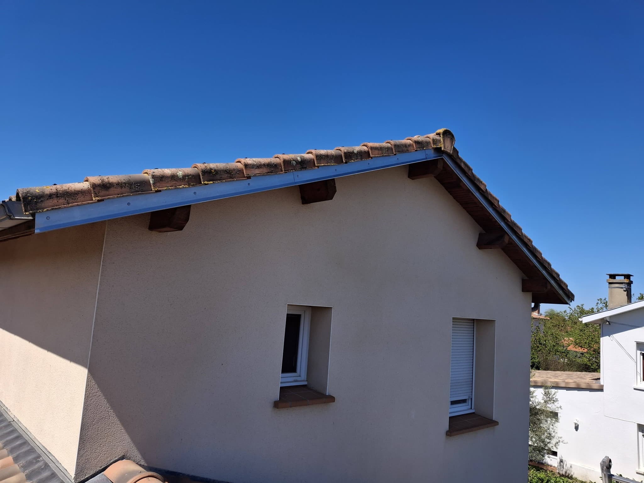 Toiture en tuiles canal sur façade beige sous un ciel bleu dégagé, avec détails de charpente et fenêtres.