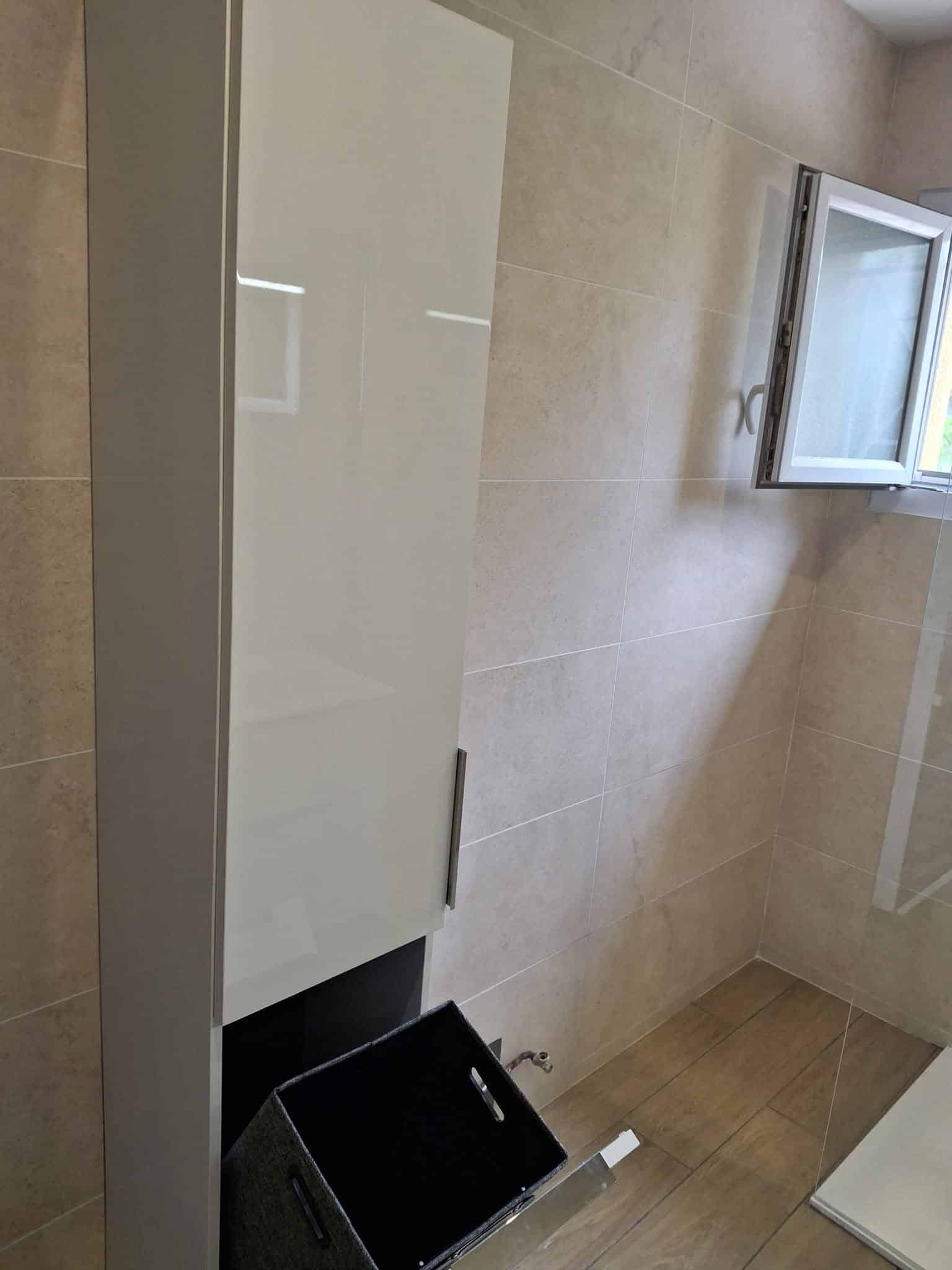 Meuble colonne blanc brillant, carrelage mural beige et fenêtre de salle de bain.
