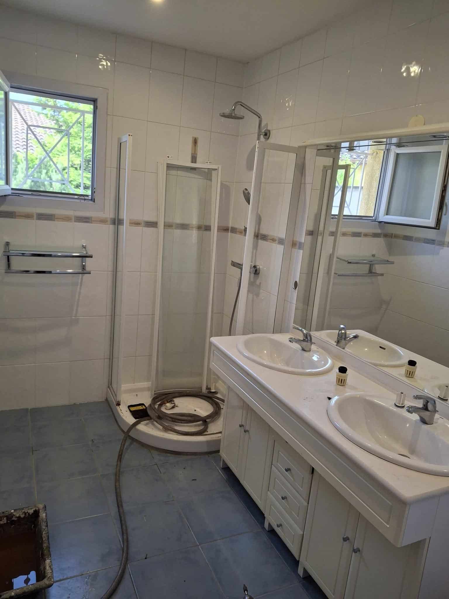 Salle de bain avec meuble double vasque blanc, douche en coin et carrelage clair.