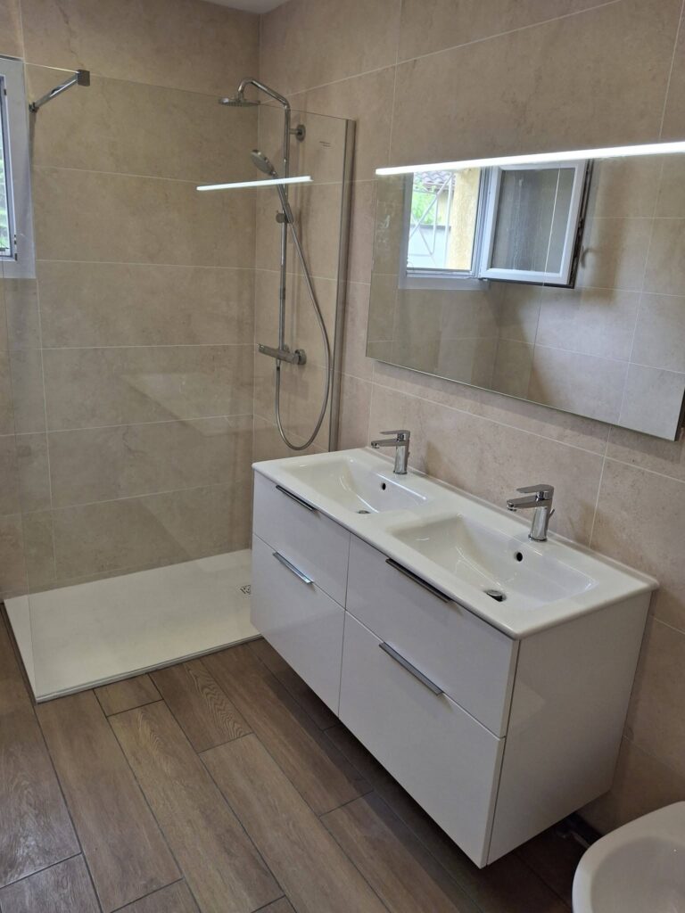 Salle de bain moderne avec douche à l'italienne, double vasque blanche et carrelage beige effet pierre.